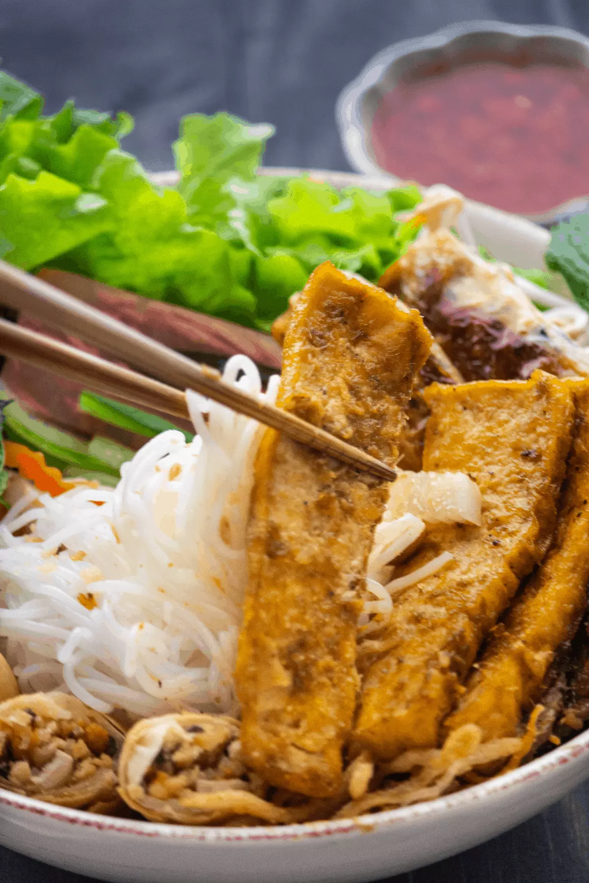 Noodles and fried tofu being held up by chopsticks in a bowl.