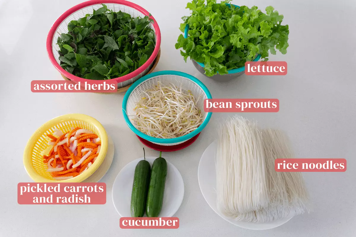 Assorted herbs in a colander next to colanders of lettuce, bean sprouts and pickled radish and carrots alongside plates of cucumbers and rice noodles.